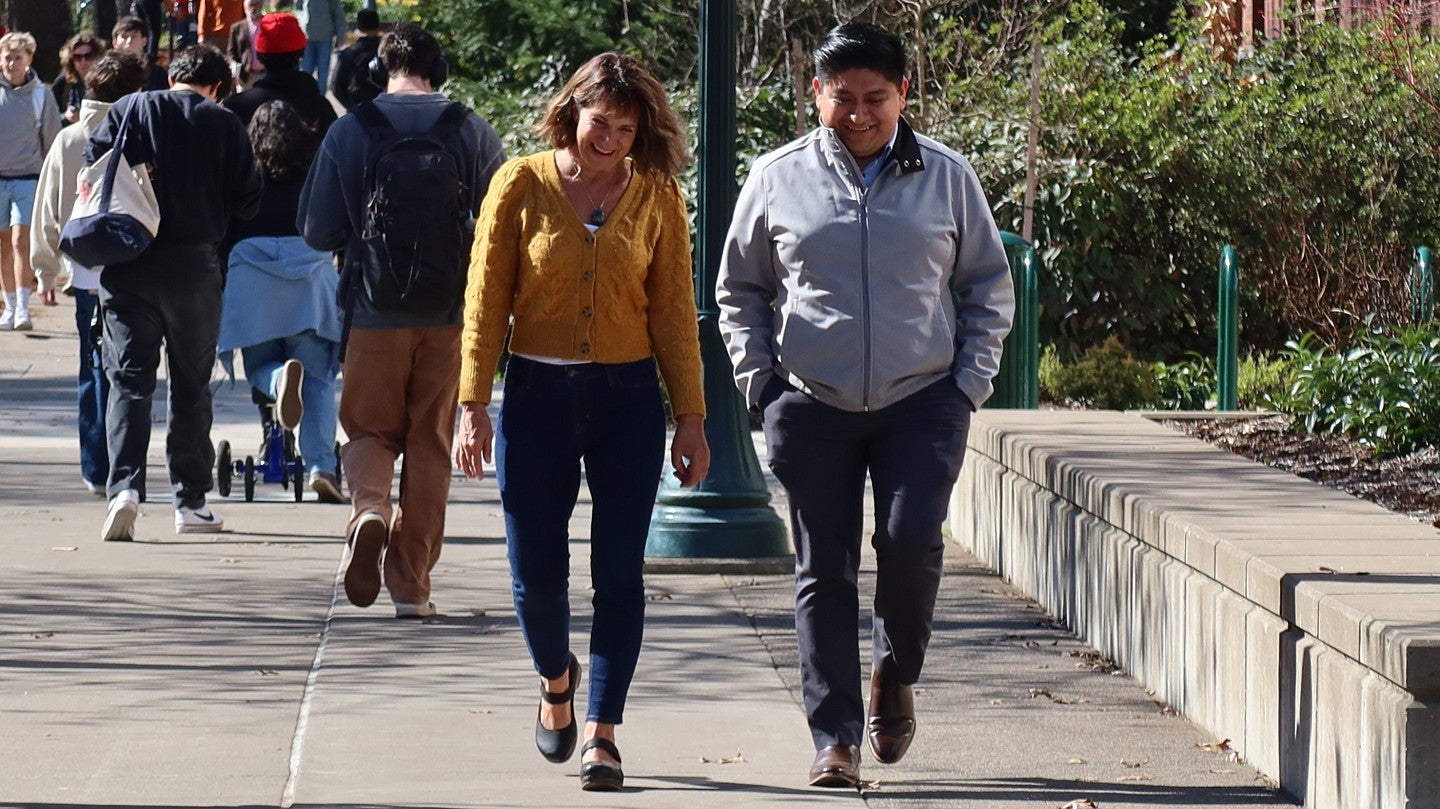 A woman and a man walk on a university sidewalk with students in the background